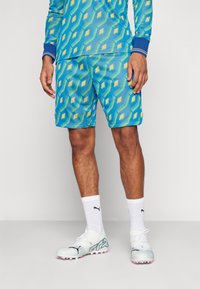Blaue, gemusterte Sportshorts mit gelben Rauten, kombiniert mit wei�ßen Socken und leichten Fußballschuhen mit pinken Akzenten.