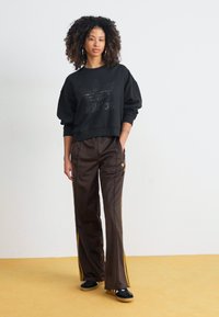 Fekete, rövidített kapucnis pulóver, dombornyomott logóval, barna, selyemhez hasonló tréningnadrággal kombinálva, amely sárga oldalsávokkal és laza szabással rendelkezik.