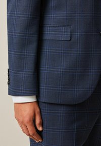 Chaqueta de traje de cuadros azul marino con un tejido texturizado que presenta líneas azules y grises. Incluye botones negros y se asoma el puño de una camisa blanca por debajo de la manga.