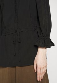 Blouse noire texturée avec une coupe décontractée, dotée d'une taille à cordon de serrage et de poignets froncés. Le matériau semble léger et légèrement transparent.