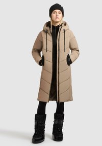 Beige, lång pufferjacka med huva, svarta detaljer och fickor med dragkedja, bärs med svarta leggings och knähöga, svarta vinterstövlar.