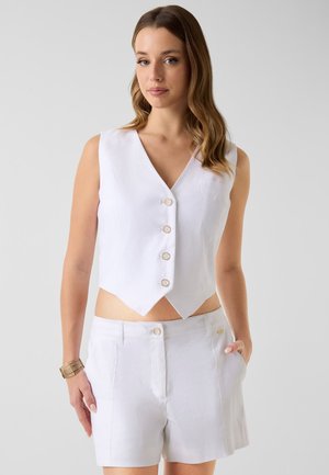 Vrouw draagt een wit mouwloos gilet met knopen en witte shorts met hoge taille, met haar rechterhand in haar zak en een gouden armband om haar linker pols.