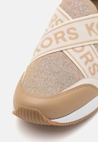 Gros plan sur l'avant d'une basket beige avec un tissu texturé, une bride croisée portant l'inscription "KORS" et une semelle blanche avec une semelle extérieure marron à motif.