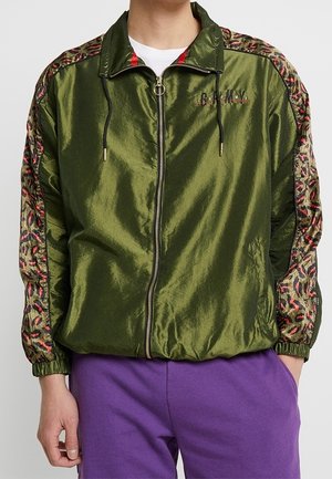 Olivgrüne Zip-Jacke mit glänzender Oberfläche, die Leopardenmuster-Akzente an den Ärmeln und der Kordelzugkapuze aufweist; gerippte Bündchen und Saum.