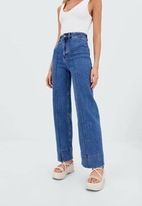 Hoge taille blauwe denim jeans met wijde pijpen, zichtbare stikdetails en een lichte textuur, gecombineerd met beige plateau sandalen.