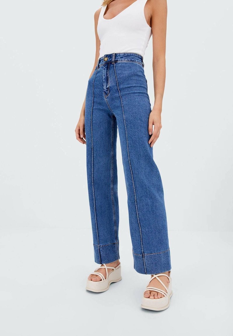 Hoge taille blauwe denim jeans met wijde pijpen, zichtbare stikdetails en een lichte textuur, gecombineerd met beige plateau sandalen.