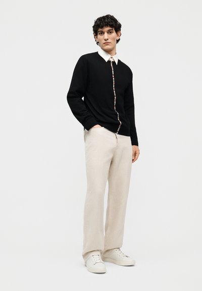 Jeune homme portant un pull noir avec un détail rayé multicolore sur une chemise à col blanc, un pantalon beige et des baskets blanches, debout avec une main dans la poche.