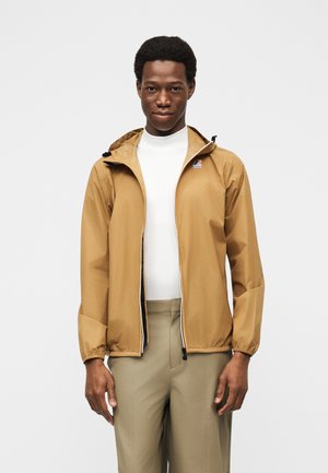 Νεαρός άνδρας με μπεζ σακάκι με κουκούλα πάνω από λευκό πουκάμισο και μπεζ παντελόνι, στέκεται μπροστά σε απλό ανοιχτό γκρι φόντο.