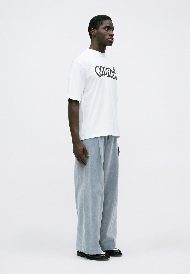 Homme se tenant de côté portant un t-shirt graphique blanc, un pantalon large bleu clair et des chaussures noires devant un fond blanc uni.