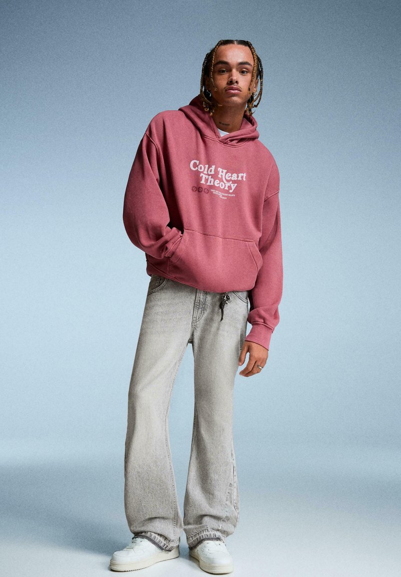 Personne avec des cheveux tressés portant un sweat à capuche rose poussiéreux avec le texte "Cold Heart Theory", un jean gris clair et des baskets blanches sur un fond gris.