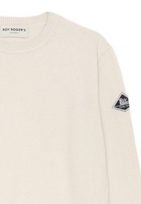 Maglione a maglia color beige chiaro con scollo rotondo e polsini a coste. Presenta una toppa a forma di rombo di colore blu scuro sulla manica sinistra con dettagli del logo.