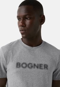 Młody mężczyzna z krótkimi włosami, ubrany w szary T-shirt z rozmytym czarnym napisem "BOGNER", patrzący w dół i w lewo na białym tle.