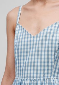 Gros plan d'une personne portant une robe sans manches à carreaux bleus et blancs avec des bretelles fines et un décolleté en V.