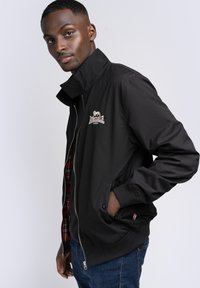 Chaqueta tipo bomber negra con un alto collar, que presenta una cremallera, bolsillos laterales y un logo bordado. La capa interior muestra un patrón de cuadros.