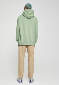 Felpa verde con cappuccio, pantaloni beige, scarpe sportive testurizzate con finitura lucida. Vista da dietro, con una vestibilità rilassata e senza modelli visibili.