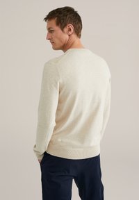 Uomo di profilo con le mani nelle tasche, indossando un maglione beige e pantaloni blu scuro su uno sfondo neutro.