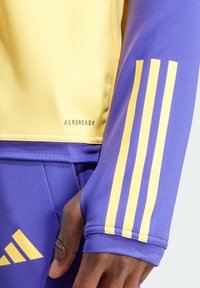 Camiseta deportiva de manga larga en púrpura y amarillo, con el texto "AEROREADY". Tres rayas amarillas en la manga púrpura, textura suave y material ligero.