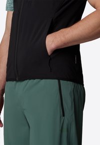 Black, sleeveless vest featuring zippered side pockets and "WINDWALL" label; paired with green shorts, textured fabric, and athletic design.