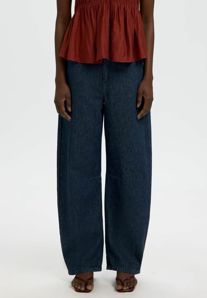 Personne portant un jean denim large et sombre, un top sans manches couleur rouille et des sandales à brides rouge foncé sur un fond blanc uni.