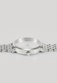 Montre en métal argenté avec un boîtier circulaire, un cadran clair, des marqueurs d'heure minimalistes et un bracelet en maille. Comprend une couronne pour les réglages.
