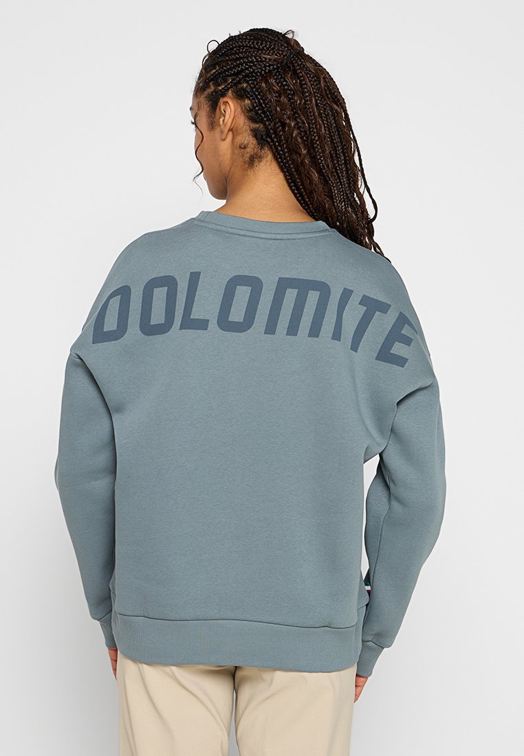 Mujer con el cabello largo trenzado, vistiendo un sudadera azul-gris con "DOLOMITE" impreso en la parte de atrás y pantalones beige, de espaldas.