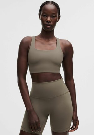 Kvinde iført taupe højtaljede biker shorts og matchende sports-bh, stående med afslappede arme mod en ensfarvet lys baggrund.