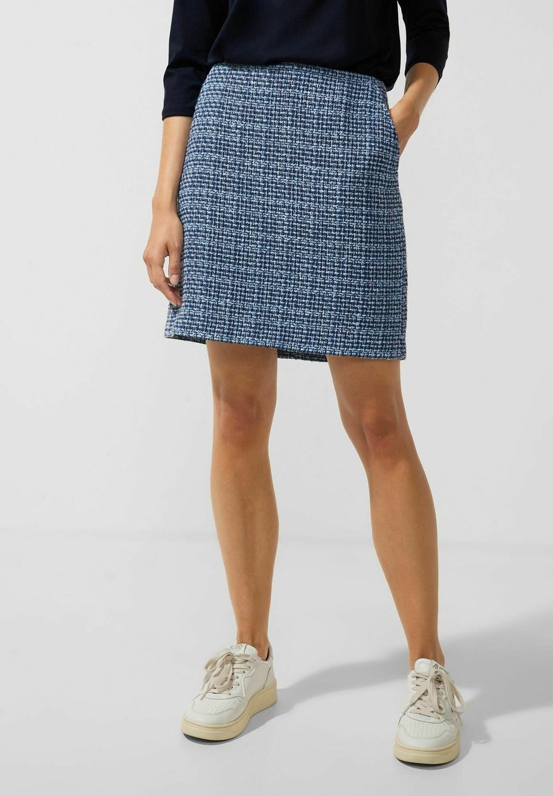 Femme portant une jupe bleue texturée avec un haut noir et des baskets blanches, se tenant avec une main dans la poche de la jupe devant un fond uni.