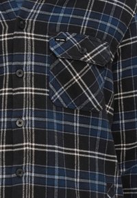Καρό πουκάμισο σε ναυτικό μπλε, μαύρο και λευκό, με κουμπιά μπροστά, μακριά μανίκια και τσέπη στο στήθος με λογότυπο. Μαλακό, υφασμάτινο υλικό με υφή.