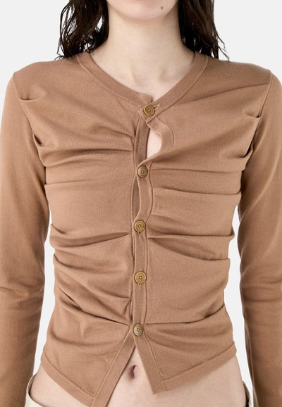 Femme portant un haut marron ajusté à manches longues boutonné avec des détails froncés et une petite ouverture en forme de goutte à la poitrine.