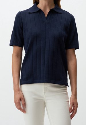 Vrouw draagt een marineblauw geribd poloshirt met korte mouwen gecombineerd met een hooggetailleerde off-white broek tegen een effen achtergrond.
