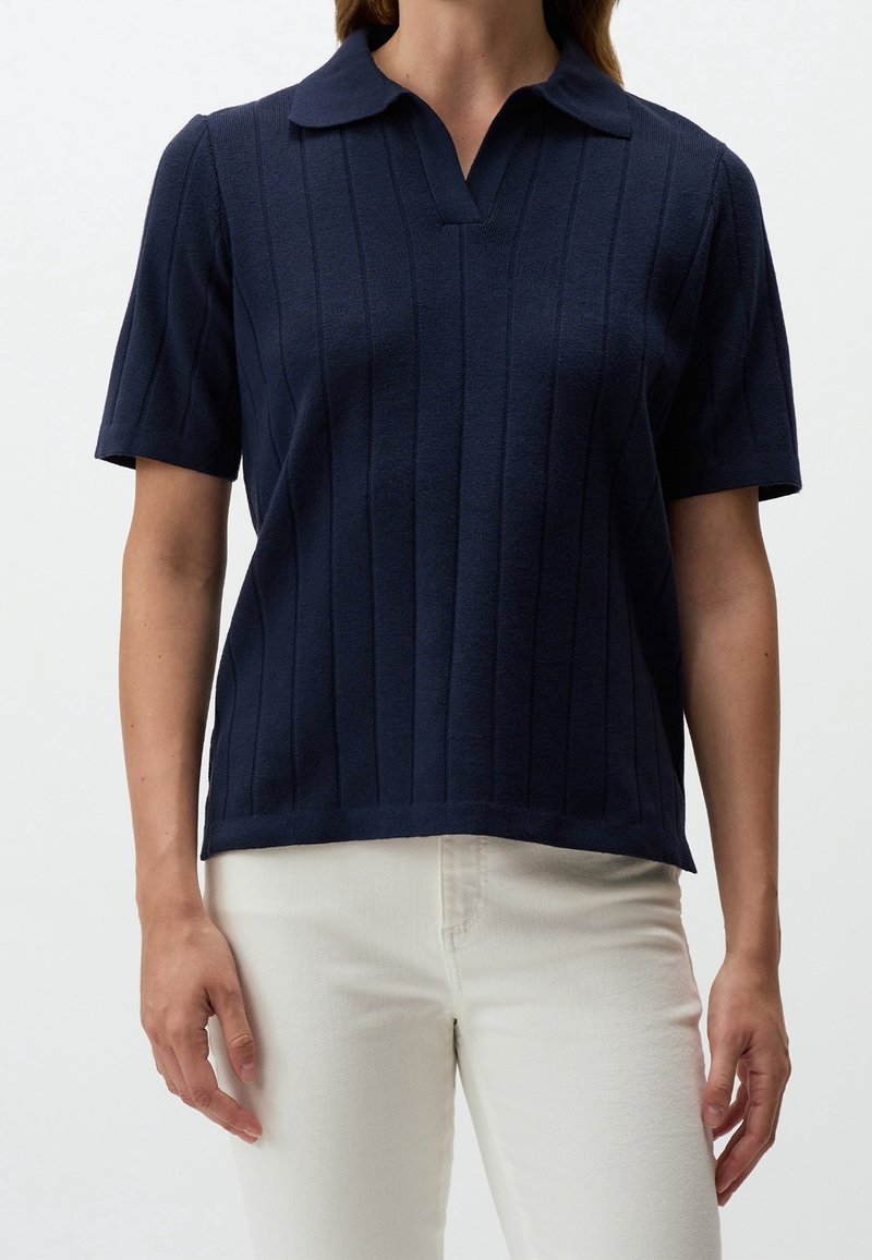 Femme portant un polo côtelé bleu marine à manches courtes accompagné d'un pantalon taille haute blanc cassé sur un fond uni.