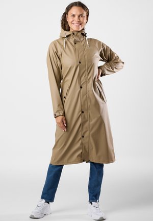 Smilende kvinde iført en lang beige regnfrakke med hætte, kombineret med blå jeans og hvide sneakers, står med den ene hånd på hoften.