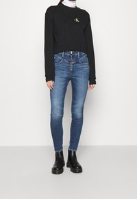 Zwart cropped sweatshirt met groen logo, gecombineerd met strakke blauwe spijkerbroek en zwarte enkellaarsjes. De spijkerbroek heeft een ritssluiting aan de voorkant en vijf zakken.