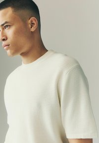 Short-sleeved, textured sweater in off-white, featuring a classic crew neckline and a woven pattern on fabric.