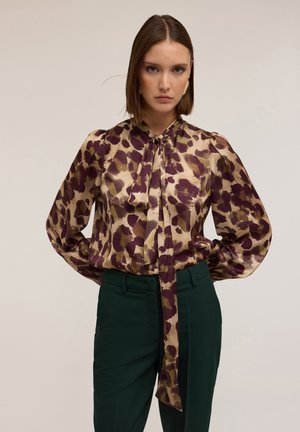 Blouse met lange mouwen van een zijdezachte stof met een bloemenpatroon in paars en beige, met een gestrikte hals en volumineuze mouwen. Gecombineerd met groene broeken.