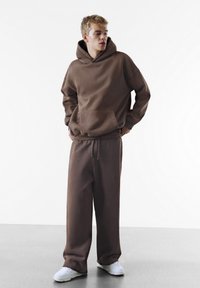 Bruine hoodie met een voorzak, gecombineerd met bijpassende bruine sweatpants. Witte sportschoenen maken de outfit compleet. Glad textuur.