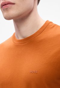 Jauns cilvēks, tērpies oranžā T-kreklā ar vārdu "BOSS", kas izcelts uz krūškurvja, uz vienkārša fona.