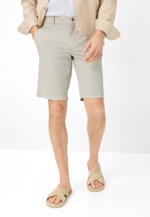 Mann, der beige Shorts, eine hellbraune Jacke, ein weißes Hemd und beige Sandalen mit Kreuzriemen trägt, steht mit einer Hand in der Tasche.