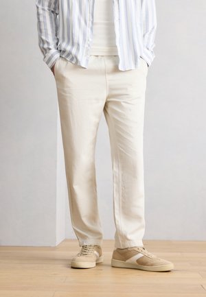 Άνδρας που φορά μπεζ παντελόνι, λευκό ριμπ πουκάμισο, γαλάζιο και λευκό ριγέ πουκάμισο με κουμπιά και μπεζ αθλητικά παπούτσια, στέκεται σε ξύλινο πάτωμα.