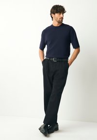 Tumši zils īss piedurkņu trikotāžas džemperis, melnas bikses ar jostu un melni apavi. Gluda tekstūra un brīvs piegriezums, stāvoša poza gaismā fonā.