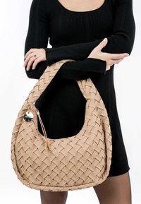 Beige woven hobo bag with a smooth texture, featuring a metallic tag and zipper, held against a black ribbed outfit.