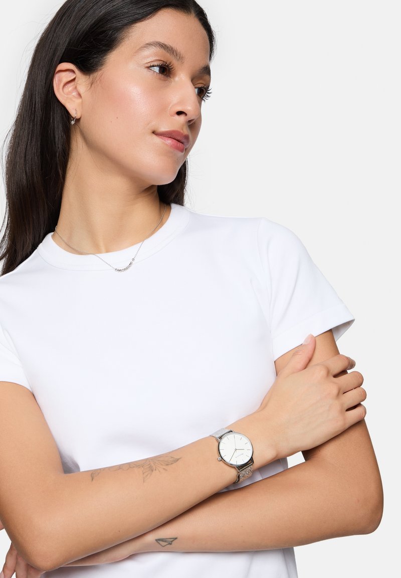 Silbernes Mesh-Armband mit einem runden weißen Ziffernblatt, getragen an einem Handgelenk mit floralem Tattoo, vor einem schlichten weißen Hintergrund und einem weißen T-Shirt.