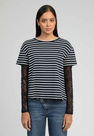 Femme aux cheveux longs et raides portant un t-shirt à manches courtes à rayures horizontales noires et blanches par-dessus un top à manches longues en dentelle noire et un jean bleu.