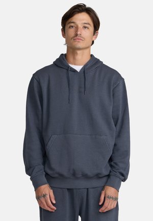 Mann trägt einen dunkelgrauen Kapuzenpullover und passende Hose mit Kordeln und Vordertasche, steht vor einem schlichten Hintergrund.