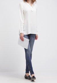 Hvid langærmet bluse med V-hals, parret med blå skinny jeans og sorte loafers; holder en hvid clutch. Enkel og moderne design.