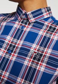 Niebieska koszula w kratę zapinana na guziki, z czerwonymi i białymi liniami, niebieskimi guzikami oraz małym logo na piersi. Wykonana z lekkiego materiału.
