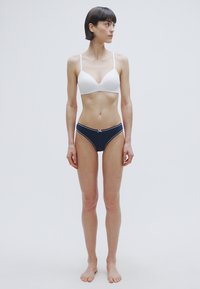 Λευκό ασύρματο σουτιέν και σκούρο μπλε μπικίνι σλιπ. Το σουτιέν έχει λεπτές τιράντες και σχέδιο με λαιμόκοψη σε σχήμα V· τα σλιπ διαθέτουν απλή αντίθετη επένδυση.