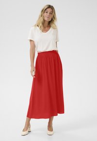 Jupe maxi rouge avec taille élastique, texture de tissu fluide, associée à un top blanc uni à manches courtes et des chaussures à talons blocs crème.