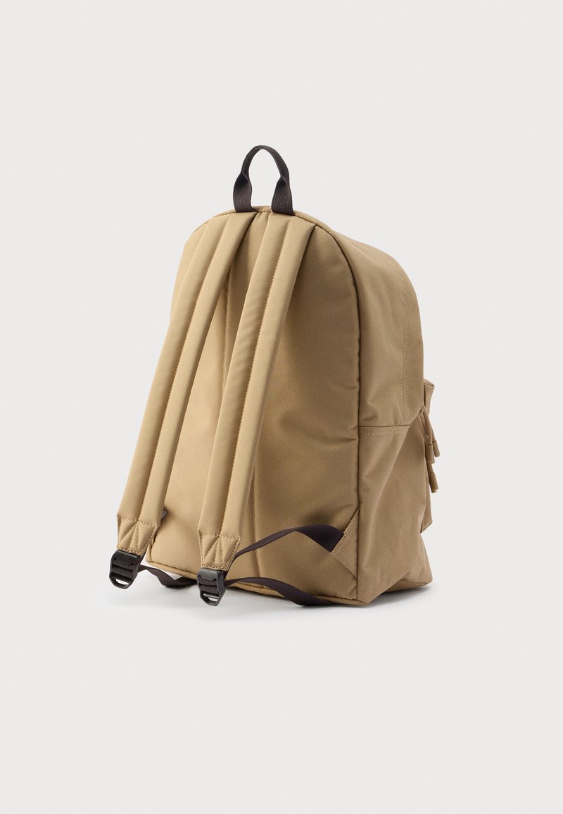 Mochila de tela beige con correas marrones ajustables, asa superior y bolsillo lateral. Textura suave y forma redondeada.