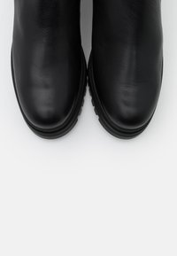 Mustat nahkasaappaat nilkkapituisella varrella, joissa on sileä pinta, pyöreä kärki ja paksu kumipohja. Yksinkertainen muotoilu ilman näkyvää metalliosaa tai kuvioita.